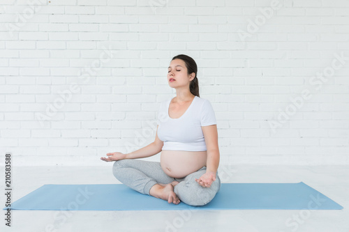 Beautiful young pregnant woman doing yoga or fitness exercise, lotus pose on the blue yoga mat at home. Happy and healthy pregnancy, meditation, calmness concept