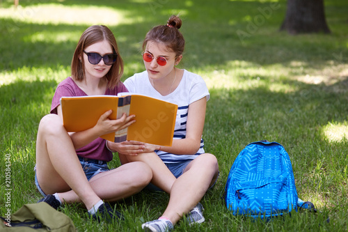Outdoor shot of pretty female students wears fashionable sunglasses, sit crossed legs on green grass in park, being focused into book, try to remember information for exam, dream about rest.