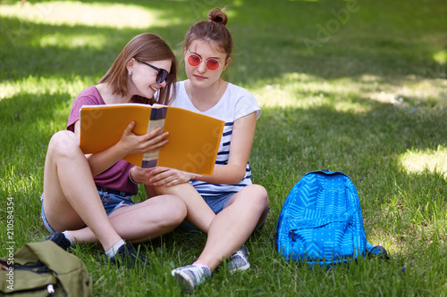 Outdoor shot of cheerful young female laughs gladfully while reads something funny in book, sits near her best female friend, dressed in fashionable clothes, rest on green grass in countryside