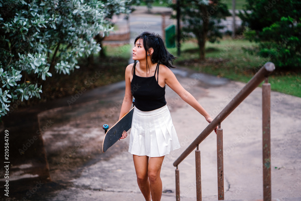 An image of a young and attractive Chinese Asian teenager wandering in the park while walking up the stairs. She is pretty, sporty, and she is carrying a skateboard with her.