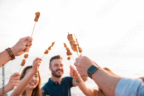 Happy friends having dinner a brochette that have been cooked in the barbecue on the beach. Summer time and holiday concept. photo