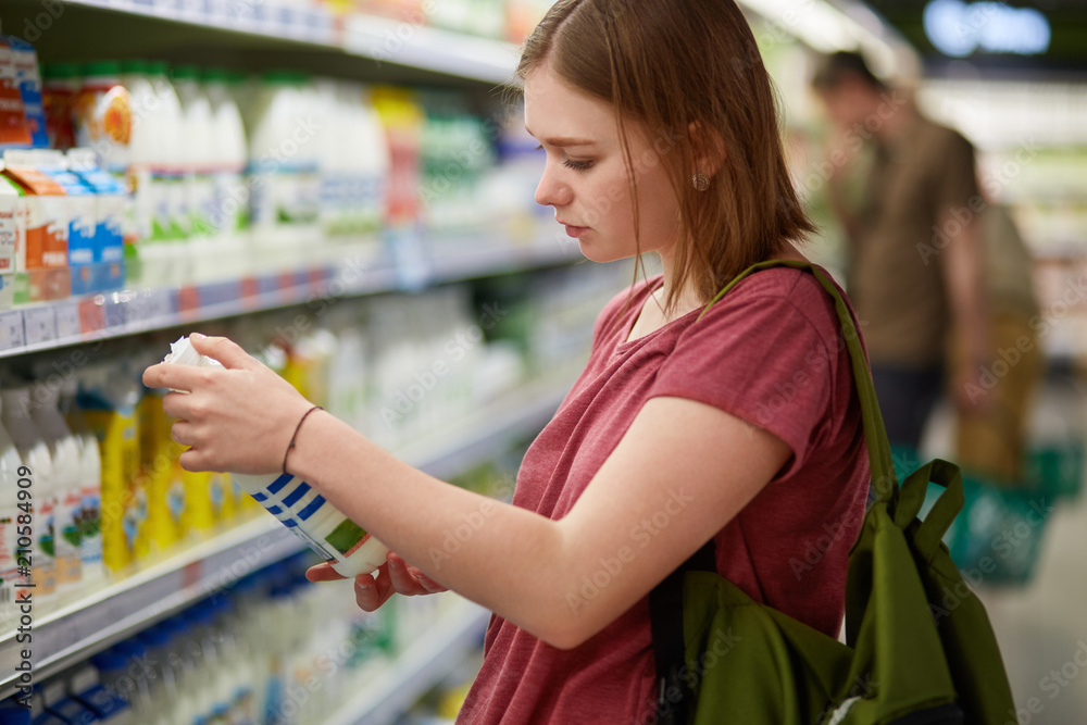 Attractive female goes shopping, stands in dairy department, holds bottle of milk, reads information on label, checks date of manufacture, carries rucksack, has serious look, stands in store