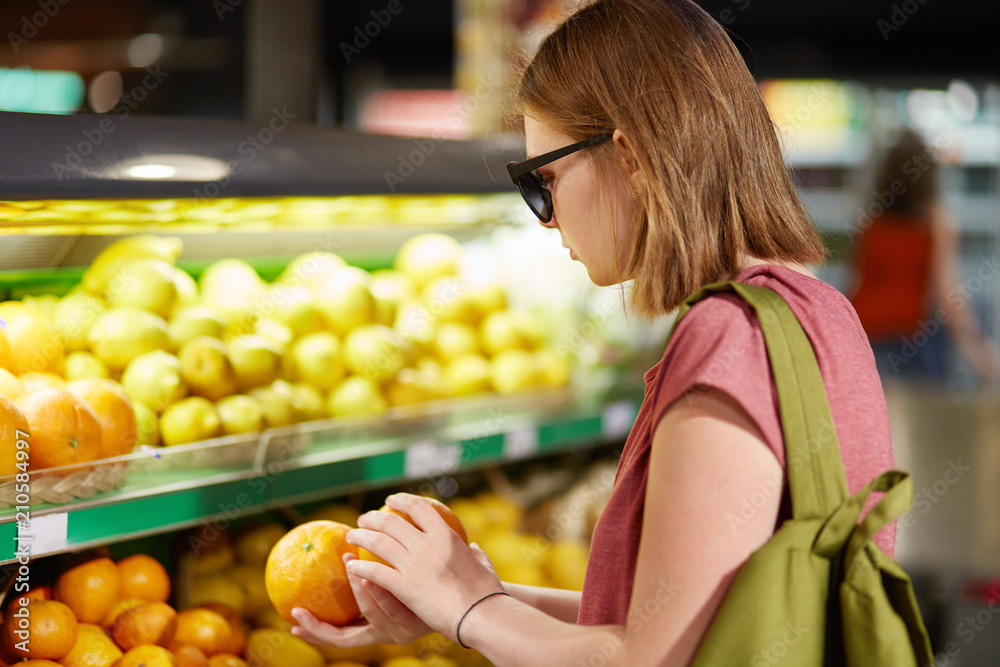 Photo of female teenager stands back, carries rucksack on back, chooses oranges for making fruit salad, wears shades, keeps to healthy diet, poses in supermerket. People and purchasing concept Photo of female teenager stands back, carries rucksack on back, chooses oranges for making fruit salad, wears shades, keeps to healthy diet, poses in supermerket. People and purchasing concept