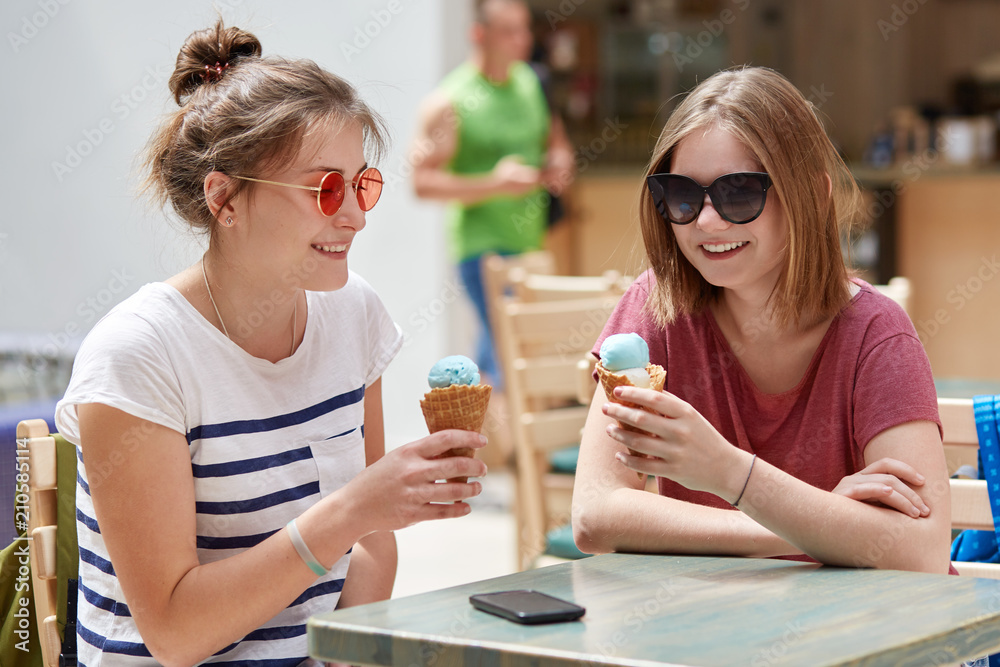 Positive female fellows have joyful expressions while recreat in cafeteria together, eat ice cream, wear sunglasses, communicate with each other, enjoy spare time during summer day. Leisure concept Positive female fellows have joyful expressions while recreat in cafeteria together, eat ice cream, wear sunglasses, communicate with each other, enjoy spare time during summer day. Leisure concept