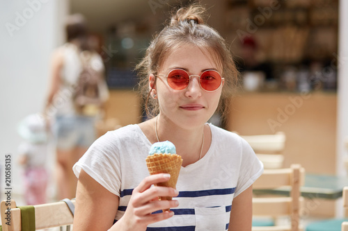 People, rest, summer, leisure concept. Pretty happy young female in red sunglasses, dressed in casual t shirt, eats delicious ice cream, enjoys vacation, strolls outdoor, has positive expression