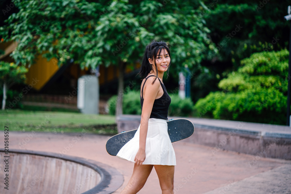 A portrait of a young, cute, and attractive Chinese Asian teenager with her skateboard in the park. She is wearing sporty attire and is smiling as she strikes a pose against the trees.
