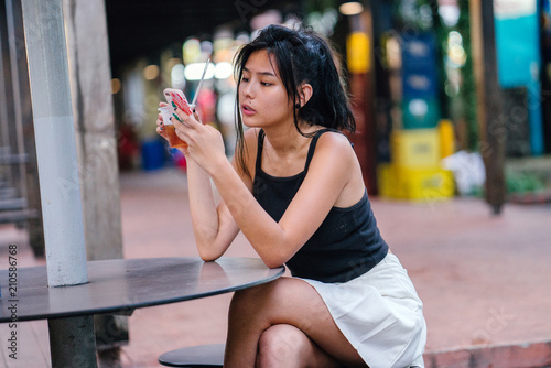 A trendy and fashionable Chinese Asian girl sitting at the hawker center while drinking her cold beverage. She is looking at her smartphone to check her social media account.