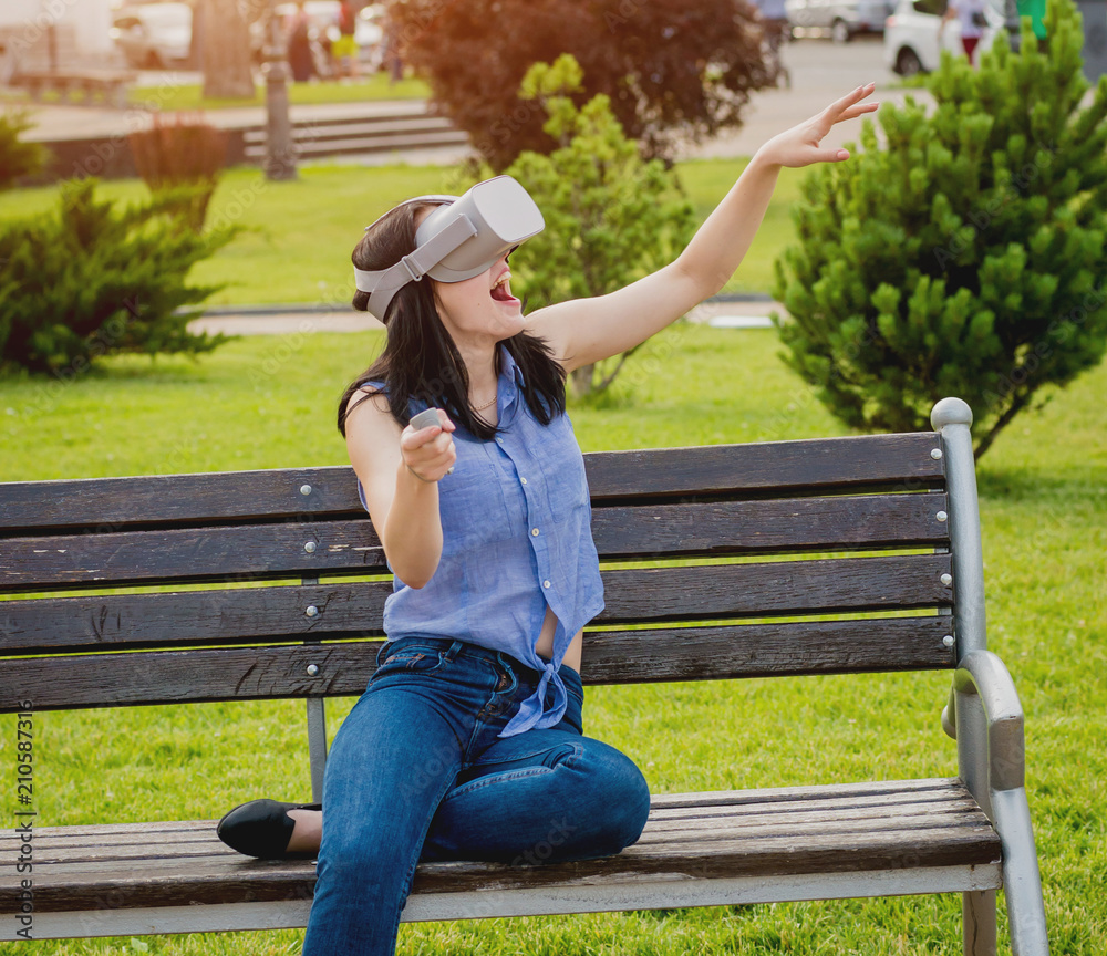 Naklejka premium A beautiful young girl plays a game wearing virtual reality glasses on the street.