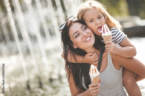 Young pretty mother and her daughter having fun together near the fountain. Beautiful woman and her little child eating ice cream. Cheerful family having fun.