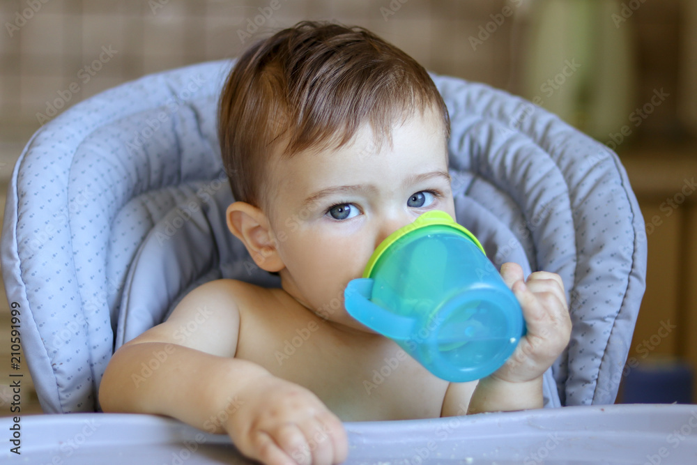 Cute blue-eyed baby boy holding plastic mug in his hands and drinking water itself lookin at camera, sitting in high feeding chair at kitchen Cute blue-eyed baby boy holding plastic mug in his hands and drinking water itself lookin at camera, sitting in high feeding chair at kitchen