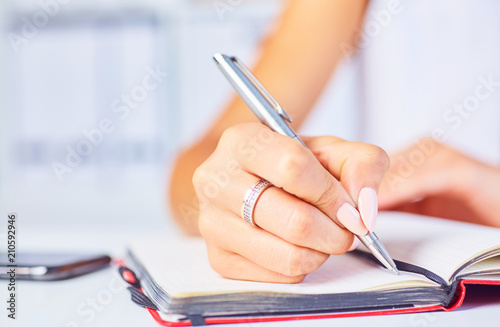 Young woman working at her desk taking notes closeup. Focus on hand writing on a notepad.
