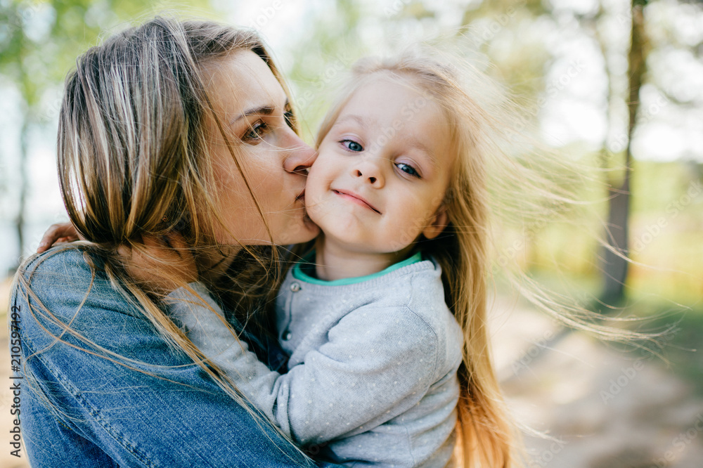 Happy smiling mother hugging her lovely little daughter outdoor. Lifestyle family. Adult cheerful female parent playing with her beautiful emotional child at nature in summer. Positive people faces. Happy smiling mother hugging her lovely little daughter outdoor. Lifestyle family. Adult cheerful female parent playing with her beautiful emotional child at nature in summer. Positive people faces.