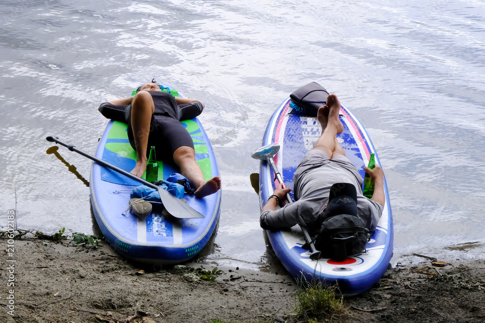 Letni wypoczynek nad wodą - kobieta i mężczyzna odpoczywający na brzegu jeziora na deskach z wiosłem, SUP, Stand Up Paddle - obrazy, fototapety, plakaty 