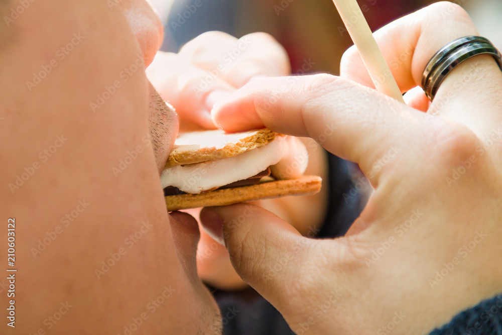 Closeup of Man Earing Smore with Roasted Marshmallow and Chocolate At Campfire Outdoors Closeup of Man Earing Smore with Roasted Marshmallow and Chocolate At Campfire Outdoors