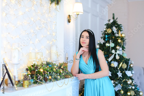 Young woman leaning against decorated fireplace, wearing blue dress and bracelet in Christmas tree background. Concept of jewelries and New Year inspiration. photo