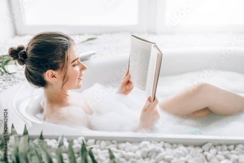 Fotografering Woman lying in bath with foam and reads magazine