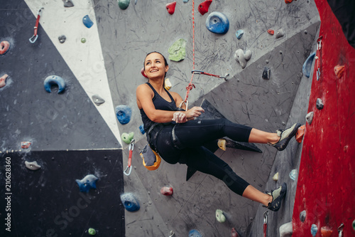 Fit sporty student girl equipped with safety rope and harness moving up at rock climbing wall at the gym. Red and grey colour background
