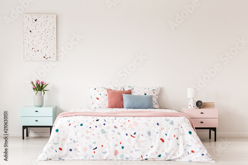 Poster above cabinet with flowers next to bed with colorful pillows in white bedroom interior. Real photo
