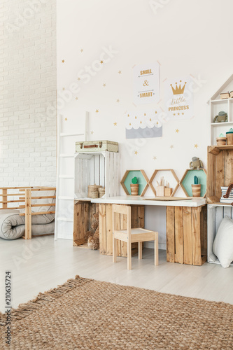 Small wooden chair standing by the desk and crate shelves in white Scandi baby girl room interior with carpet and posters on the wall