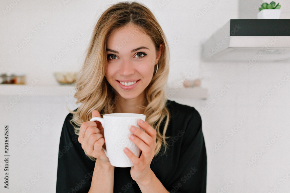 Happy beautiful caucasian blond woman in sleepwear drinks coffee in the kitchen and smiles at camera at home woke up in the morning on a sunny day. Happy beautiful caucasian blond woman in sleepwear drinks coffee in the kitchen and smiles at camera at home woke up in the morning on a sunny day.