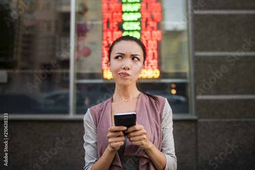 Fotografi A young beautiful asian woman using an application in her smart phone to check currency exchange rates in front of an illuminated information board