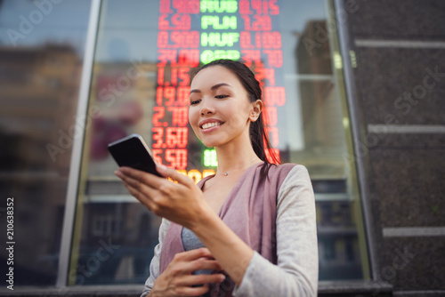 Fotografi A young beautiful asian woman using an application in her smart phone to check currency exchange rates in front of an illuminated information board