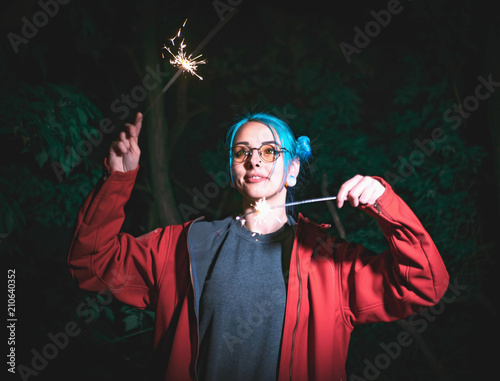 Portrait of hipster girl with blue dyed hair with sparklers in hands and reflection of it in glasses. Close up beautiful young teenage standing at night city, having fun with bengal fire. photo