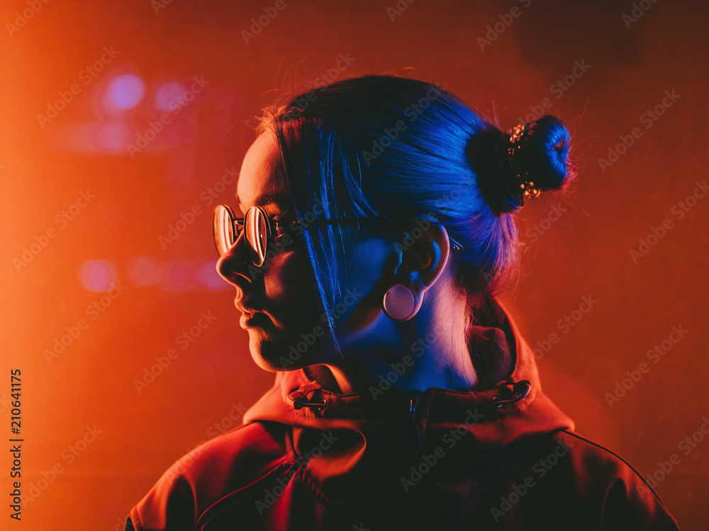 Millennial pretty girl with unusual hairstyle near glowing red neon of city at night. Dyed blue hair in braids. Mysterious hipster teenager in glasses. Reflection of light. - obrazy, fototapety, plakaty 