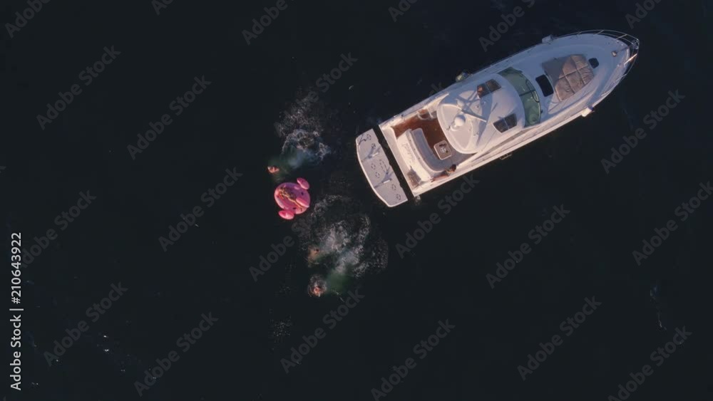 Aerial shot of friends diving in the sea from yacht and getting on a inflatable mattress. Group of friends enjoying a summer day sailing and swimming in ocean.
