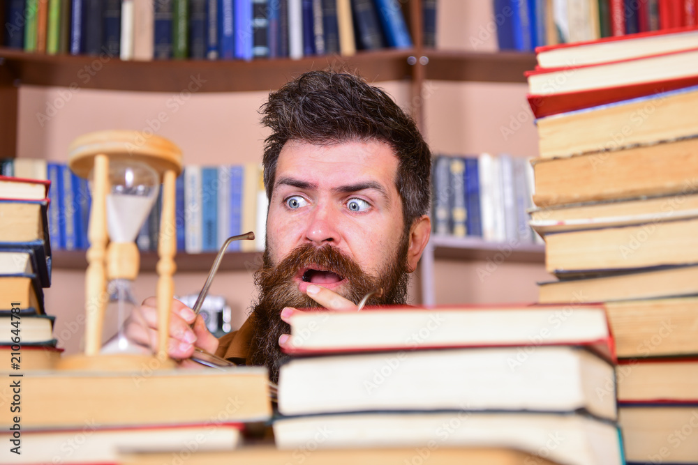 Man, scientist looks at hourglass. Teacher or student with beard studying in library. Man on serious and shocked face watching time is going over, bookshelves on background. Time flow concept. Man, scientist looks at hourglass. Teacher or student with beard studying in library. Man on serious and shocked face watching time is going over, bookshelves on background. Time flow concept.