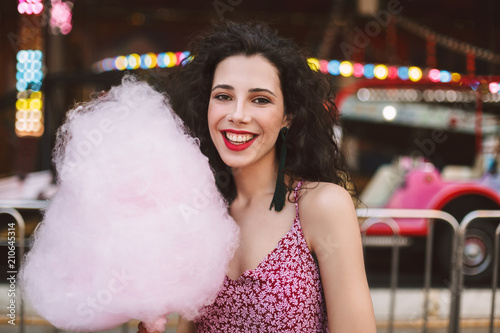 Portrait of smiling lady with dark curly hair in dress standing with cotton candy in hand and happily looking in camera while spending time in amusement park