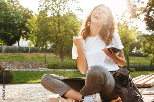 Bild auf Leinwand Photo of happy sunlit woman screaming and rejoicing while sitting on bench in gr