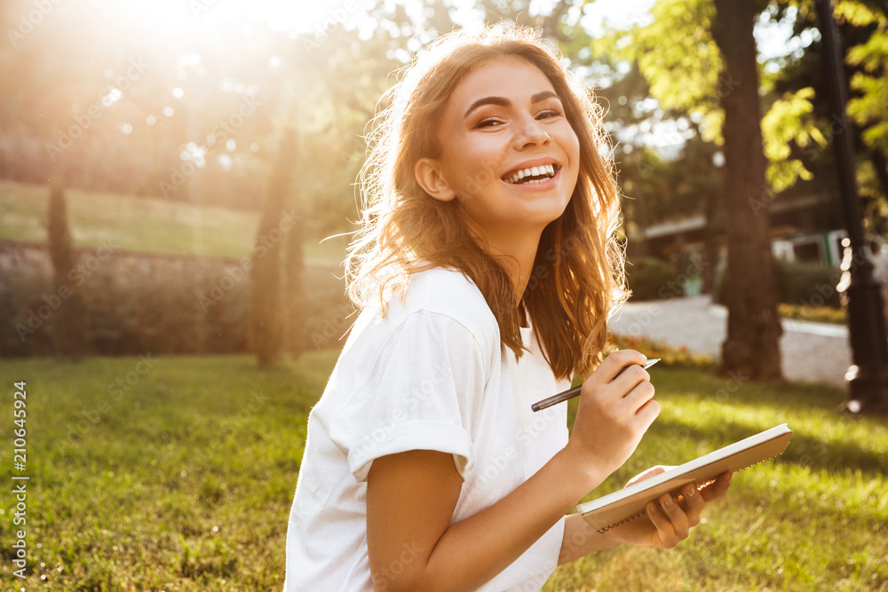 Obraz premium Zdjęcie radosnej kobiety po dwudziestce z szerokim uśmiechem siedzącej na zielonej trawie w parku ze skrzyżowanymi nogami w letni dzień i zapisującej notatki piórem