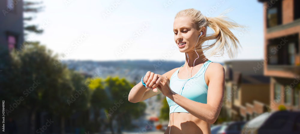 sport, technology and healthy lifestyle concept - smiling young woman with fitness tracker and earphones exercising over san francisco city background