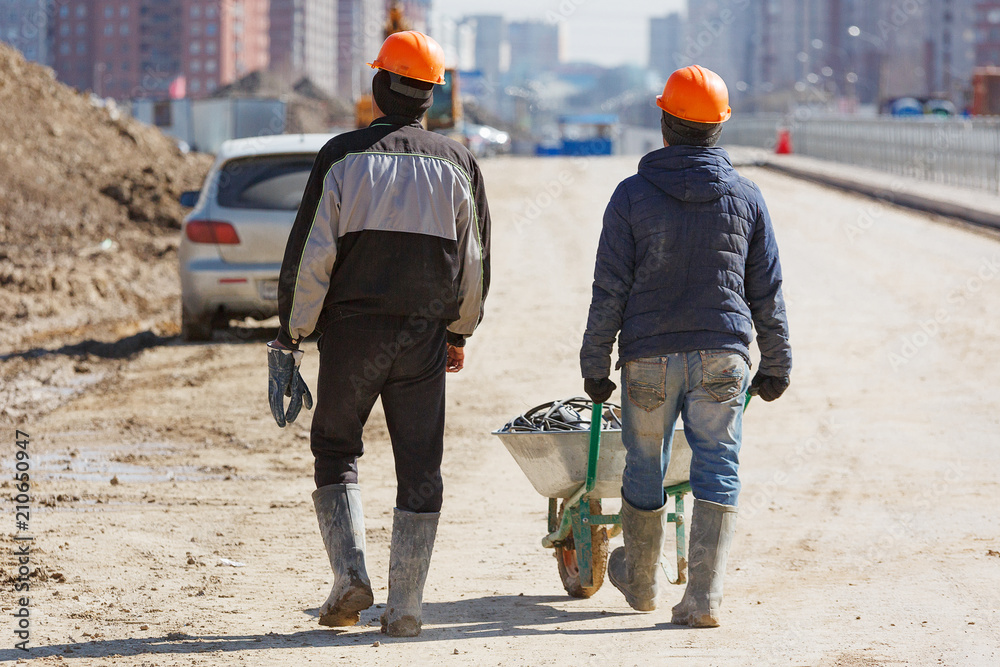 Two workers in helmets drive a wheelbarrow against the background of new buildings. Two workers in helmets drive a wheelbarrow against the background of new buildings.