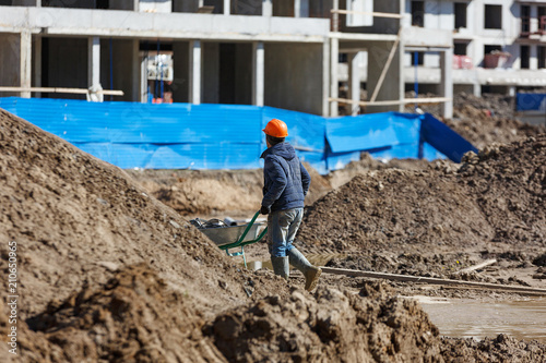 A worker in a helmet carries a wheelbarrow against the background of construction debris.