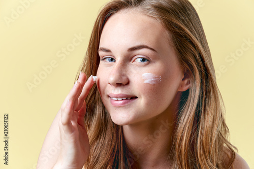 Young attractive redhead woman applying moisturiser face cream, smiling and looking at camera. Beauty, skincare, puberty concept.