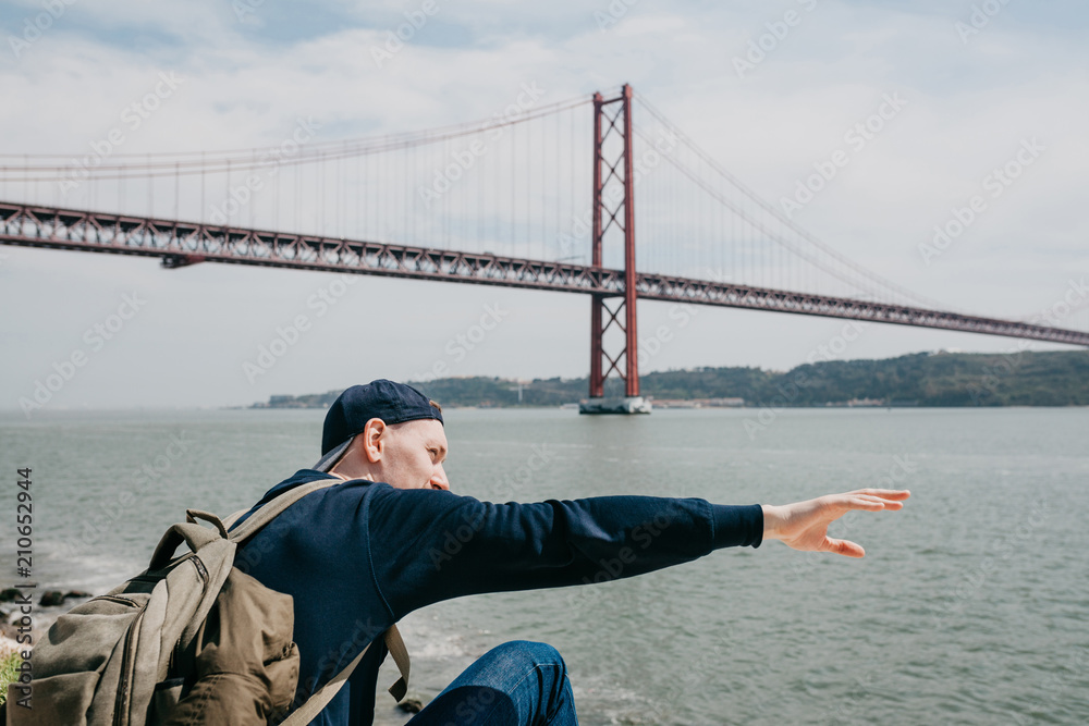 Naklejka premium A young traveler or a tourist with a backpack on the waterfront in Lisbon in Portugal next to the 25th of April Bridge