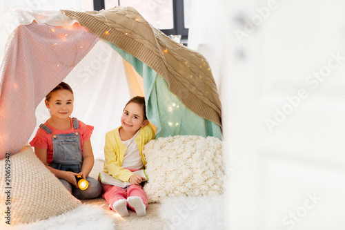 childhood, friendship and hygge concept - happy little girls with book and torch light in kids tent or teepee at home photo