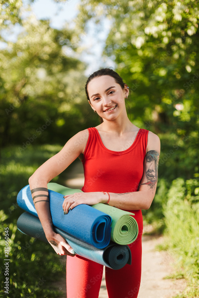 Young smiling lady in red jumpsuit standing with yoga mats in hands and joyfully looking in camera in park Young smiling lady in red jumpsuit standing with yoga mats in hands and joyfully looking in camera in park
