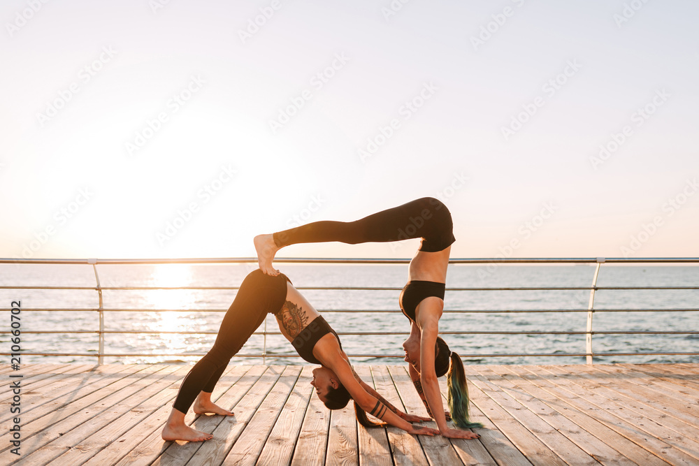 Two young ladies in black sporty tops and leggings standing and training yoga poses by the sea. Pretty women practicing yoga together with beautiful sea view on background
