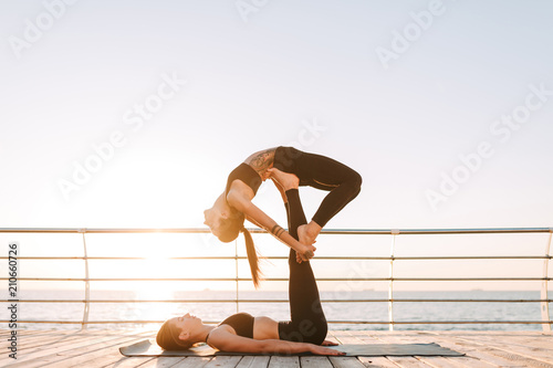 Two cool ladies in black sporty tops and leggings training yoga poses by the sea. Young women practicing yoga together with beautiful sea view on background