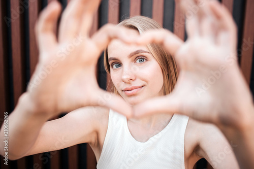 Portrait of a girl making a heart from her fingers and showing it on camera. Also she is looking at it and smiling. Isolated on striped background.