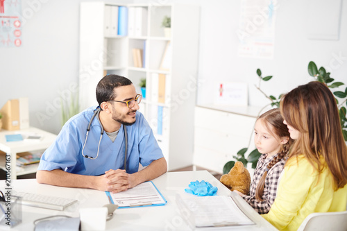 Portrait of smiling Middle-Eastern pediatrician talking to young mother and little girl during consultation in modern clinic, copy space