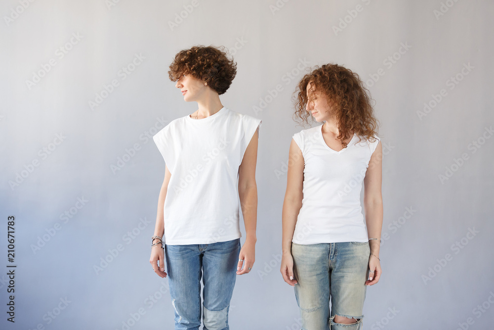 Studio shot of two trendy stylish hipster curly brunette girl, stand on concrete wall background, they turned their heads the same way. Young caucasian twin sisters wearing white t-shirt and jeans Studio shot of two trendy stylish hipster curly brunette girl, stand on concrete wall background, they turned their heads the same way. Young caucasian twin sisters wearing white t-shirt and jeans