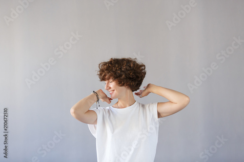 Сoncept dizzying success. Young wonderful girl keeps herself for curly head, rejoices in her youth, has great good mood. Copy space, gray studio background. Happy joyful female student smiling