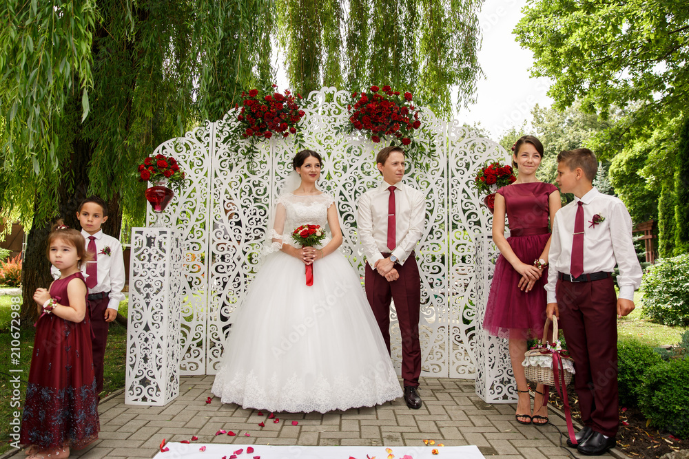 Naklejka premium Young gorgeous bride and handsome groom standing near the wedding ornament wall, with bridesmaid, groomsman and kids, wedding ceremony. At the park. Marsala style.
