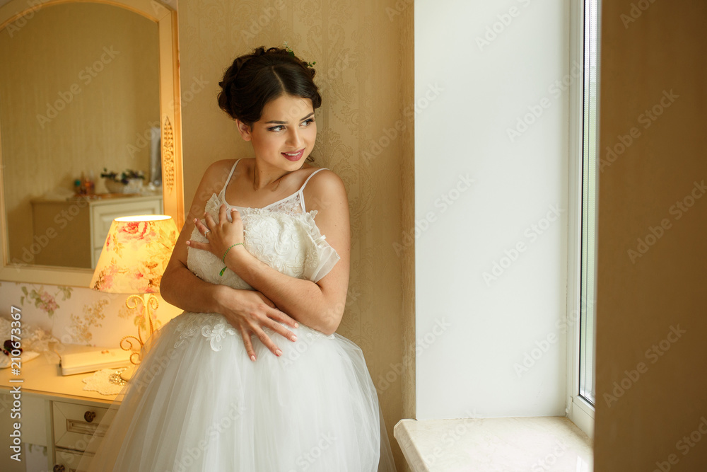 Gorgeous smiling bride holding her white wedding dress standing near the window and looking out of it. Indoors. Gorgeous smiling bride holding her white wedding dress standing near the window and looking out of it. Indoors.