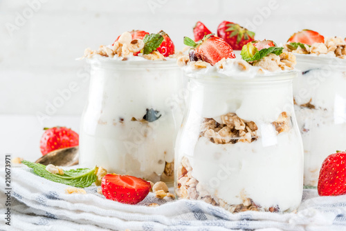 Healthy summer breakfast idea, homemade layered parfe dessert in small jar with granola, yoghurt and strawberry, dark background copy space photo