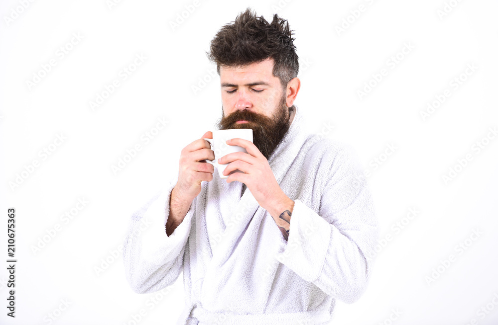 Macho drowsy, sleepy face drinks coffee in morning enjoying aroma. Man with beard and disheveled hair stands in bathrobe, holds mug with tea or coffee, white background. Morning rituals concept.